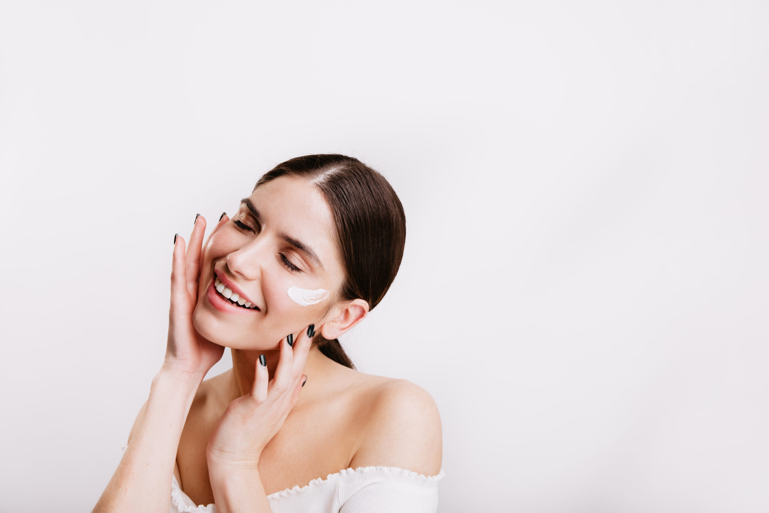 Adult woman in white top cares for skin of face, applying cream. Portrait of girl with ponytail on isolated background