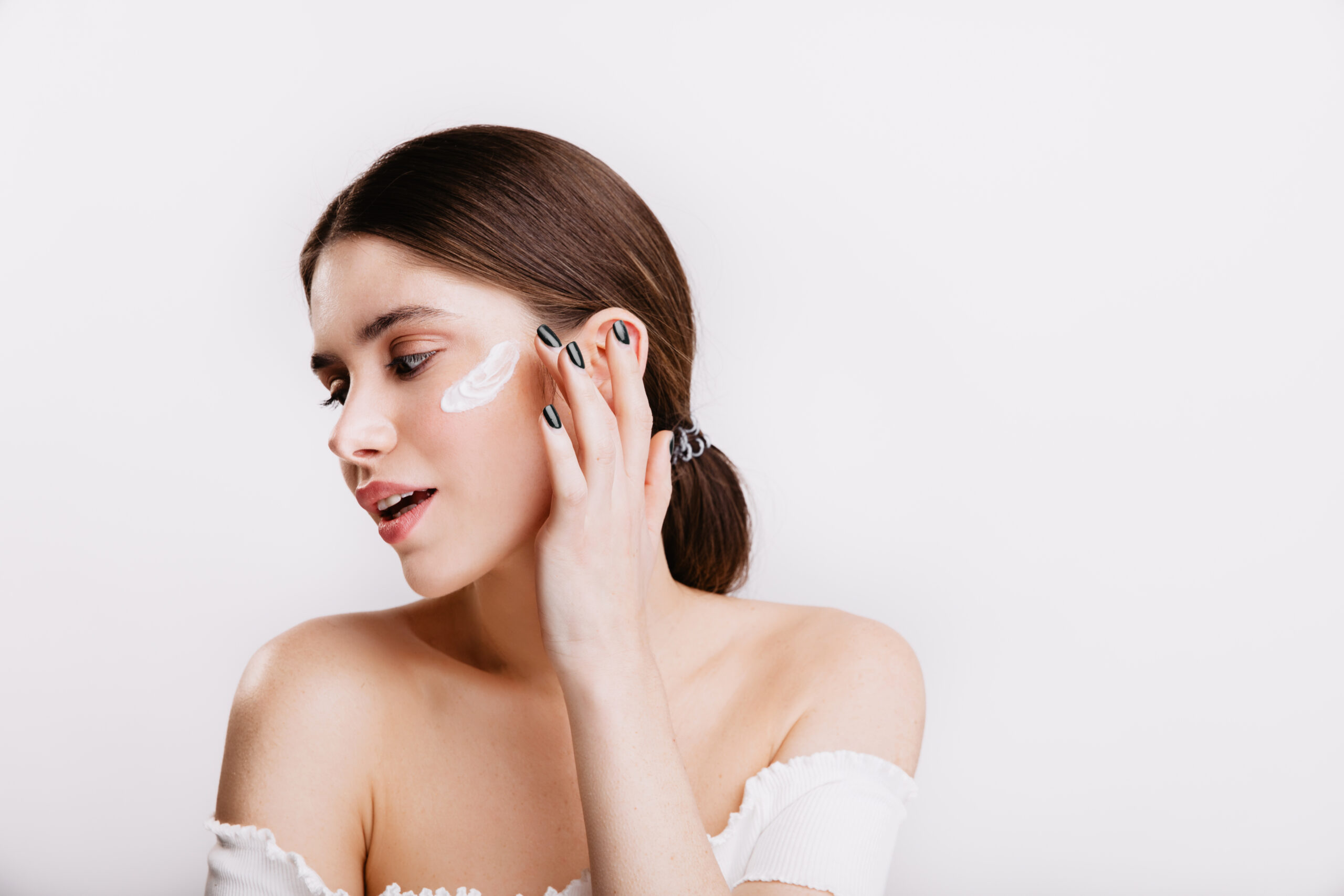 Dark-haired lady without makeup puts moisturizer on her face. Girl with black manicure gently touches skin